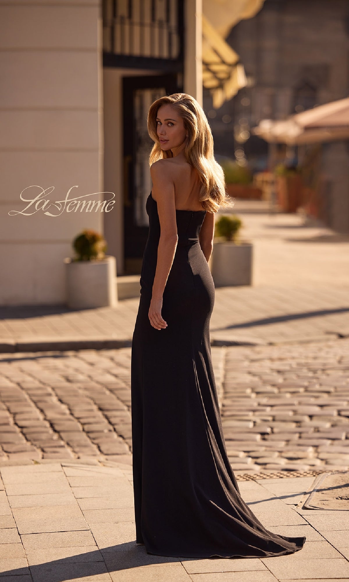 A woman in the La Femme 32919 Strapless Sweetheart Prom Dress stands outdoors on a sunlit cobblestone street, gazing back over her shoulder.